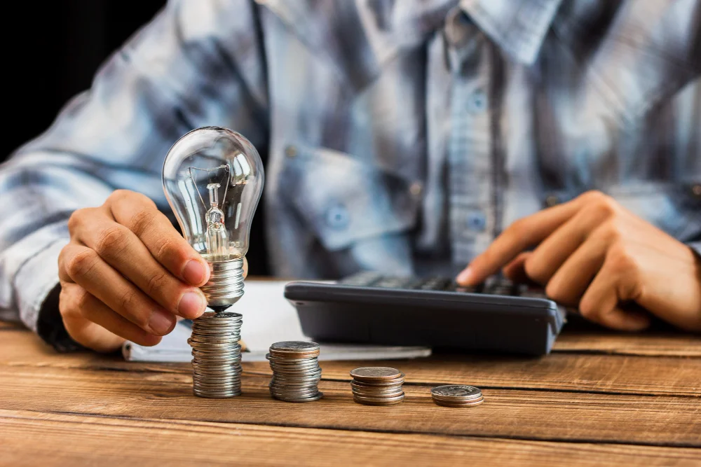 Pessoa segurando lâmpada sobre pilhas de moedas enquanto utiliza calculadora simbolizando planejamento financeiro no microcrédito para desenvolvimento econômico.